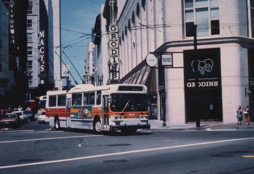 A white and orange bus, number 5044, is driving down a city street on a sunny day. Storefronts for Grodins and Macy's are visible, along with overhead wires and a few pedestrians on the sidewalk.