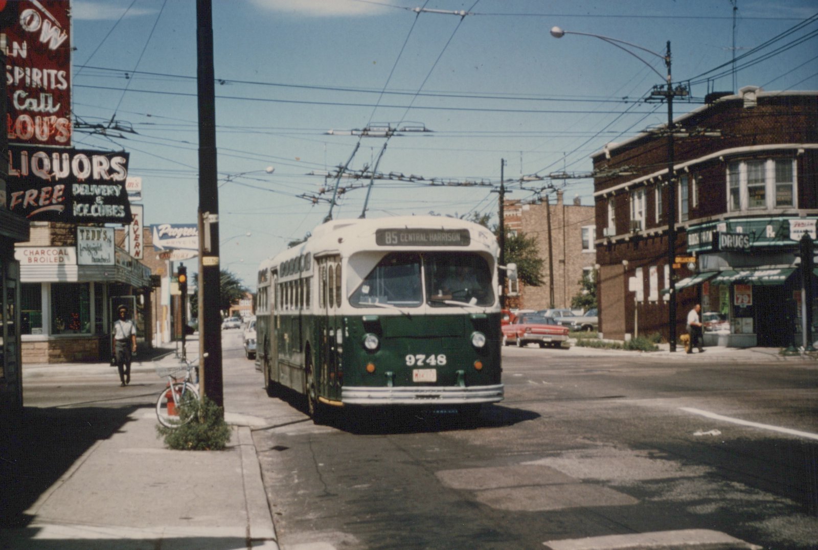 A green and cream trolleybus labeled 9748 and route 85 Central-Harrison is turning a corner on a sunny street in Chicago, with storefronts and pedestrians nearby.