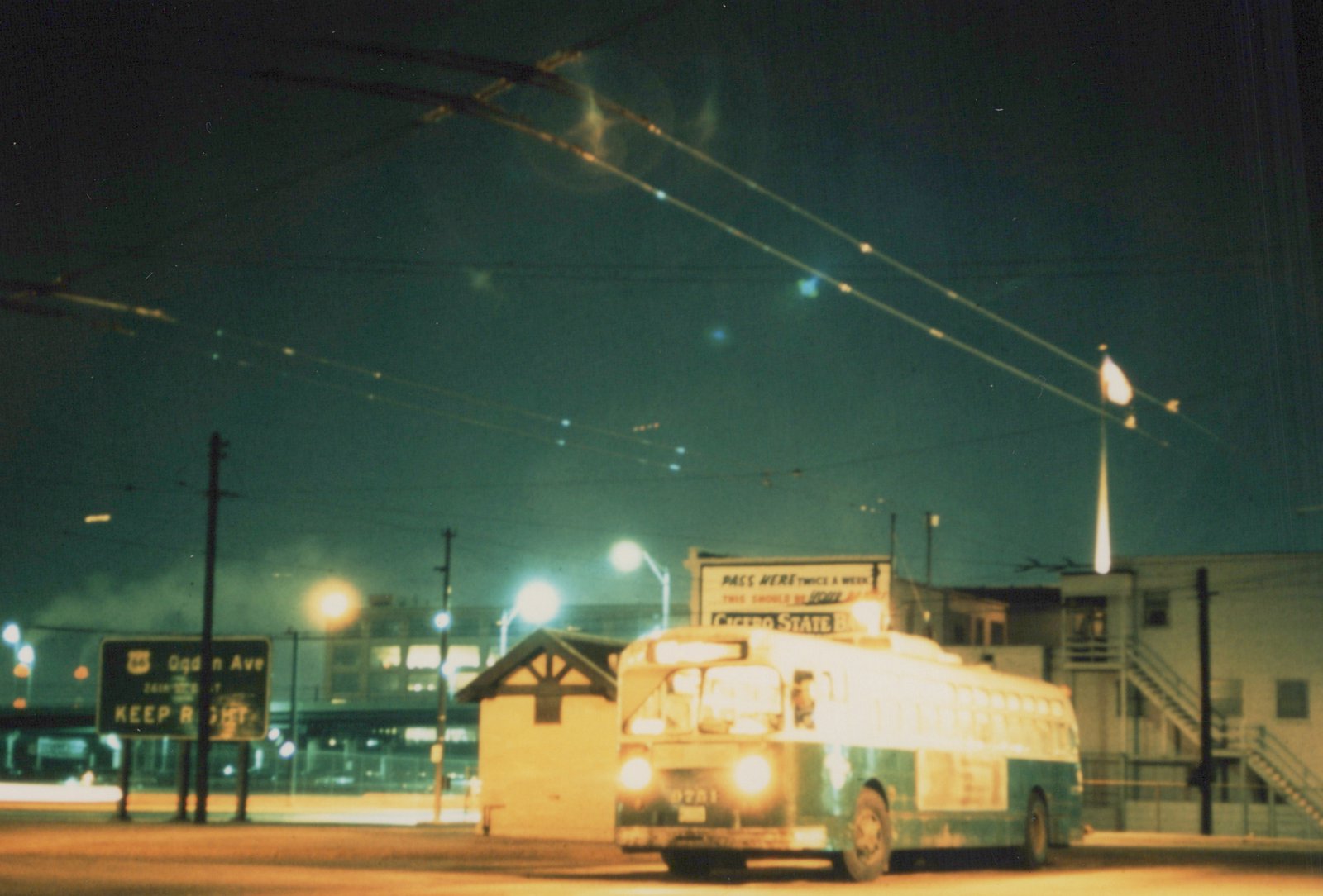 A green and cream CTA bus drives down a city street at night, headlights on, with a sign for Ogden Avenue and overhead wires visible.