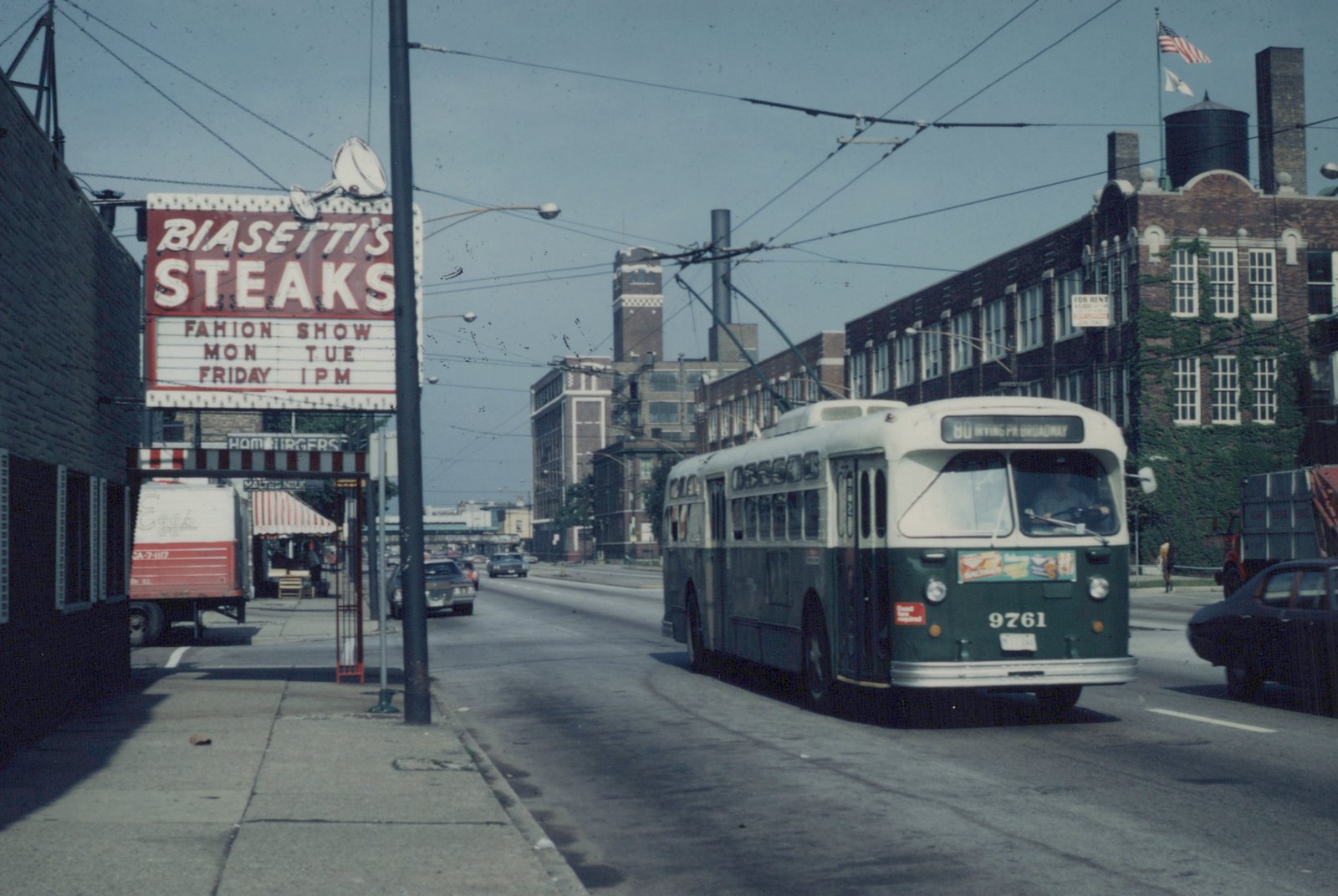 A green and cream trolleybus with the number 9761 and destination '80 IRVING PK BROADWAY' travels down a city street under overhead wires, with a 'BIASETTI'S STEAKS' sign visible on the left and brick buildings in the background.