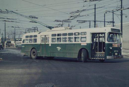 A green and cream CTA trolleybus numbered 9739, showing its destination as Irving Park, parked on a wet street under a web of trolley wires.