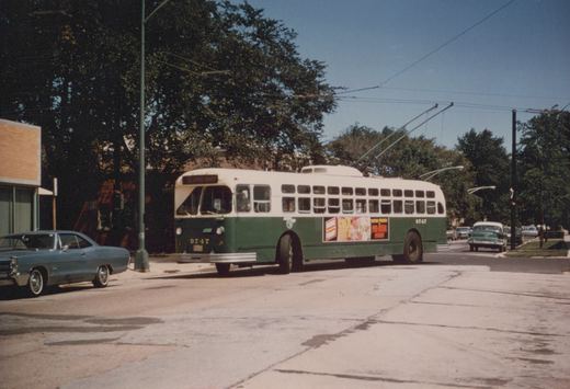 A green and cream Chicago Transit Authority trolleybus, number 9747, is driving down a tree-lined street on a clear day, with a blue car parked on the left and other vehicles in the background.