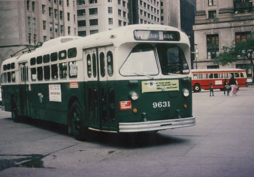 A green and white Chicago Transit Authority trolleybus, numbered 9631, is parked on a city street with its destination sign reading '77 BELMONT-CENTRAL'. In the background, a red and cream CTA diesel bus is visible along with a few pedestrians.