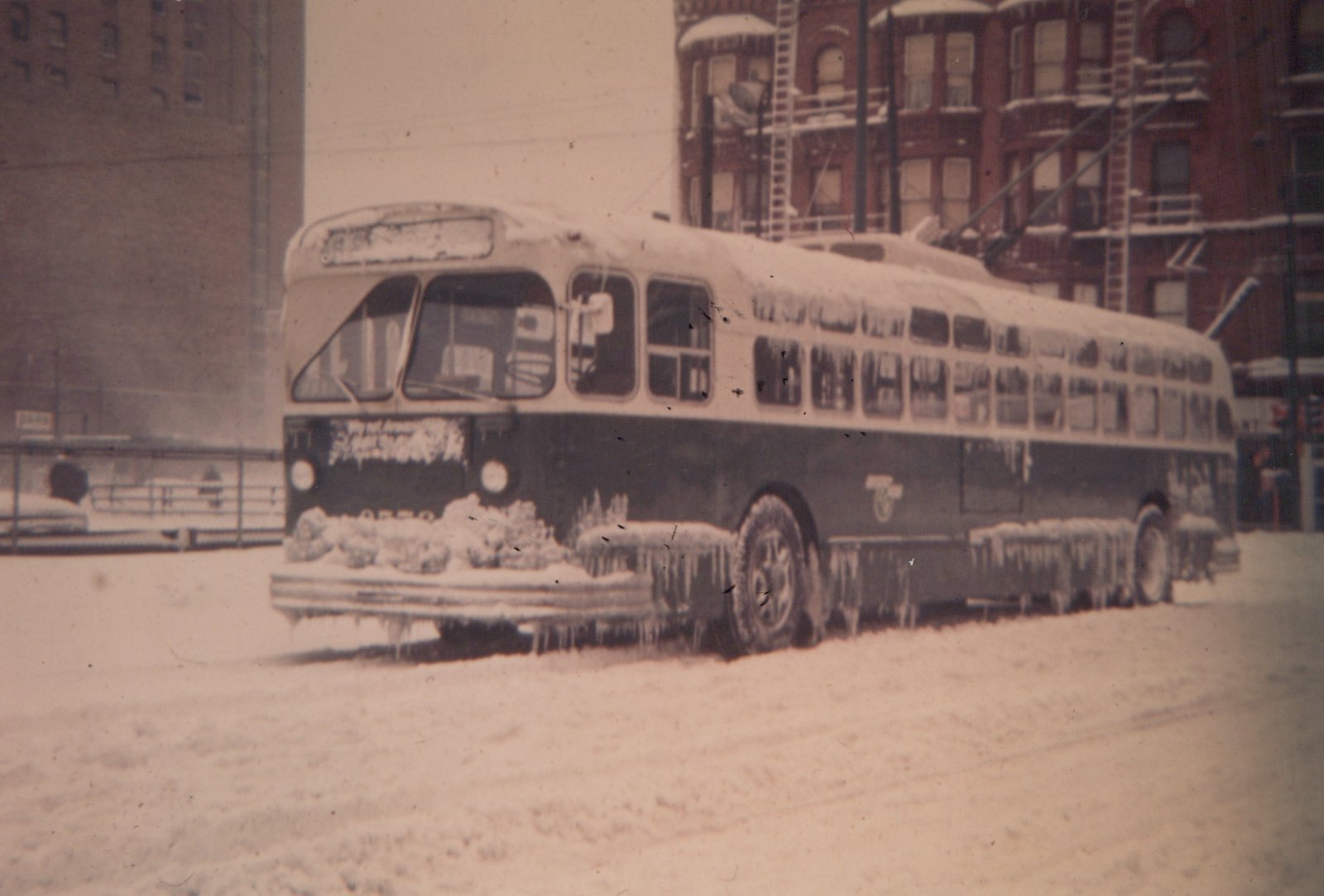 A vintage trolleybus covered in snow and ice, parked on a snowy street with snow-covered buildings in the background.