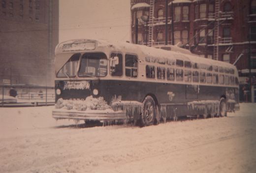 A vintage trolleybus covered in snow and ice, parked on a snowy street with snow-covered buildings in the background.