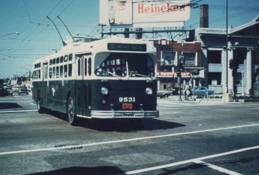 A green and cream trolleybus with the number 9531 on its front is driving down a city street with overhead wires, past a Heineken billboard and a restaurant, with several cars and pedestrians in the background.