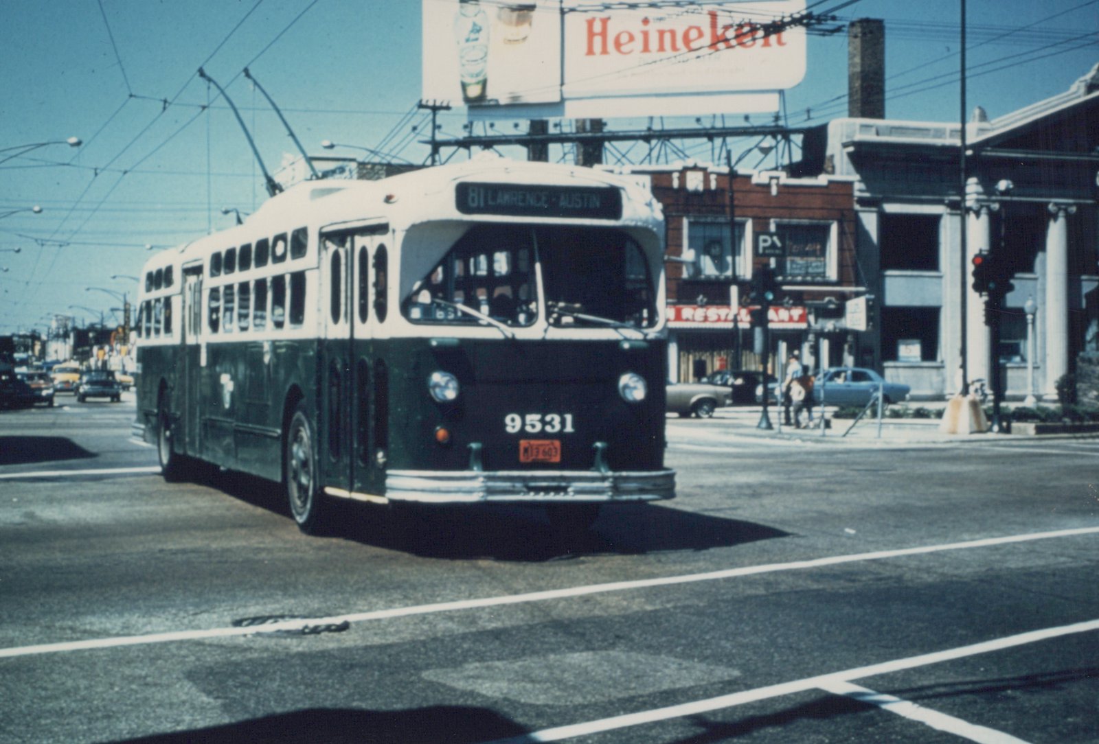 A green and cream trolleybus with the number 9531 on its front is driving down a city street with overhead wires, past a Heineken billboard and a restaurant, with several cars and pedestrians in the background.