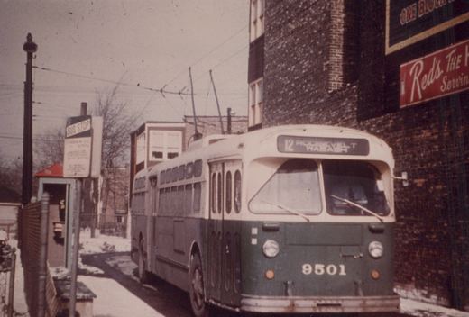 A green and cream trolleybus with the number 9501 and route 12 to Roosevelt and Wabash is parked at a snowy street corner next to a brick building with a sign for 'Red's.'