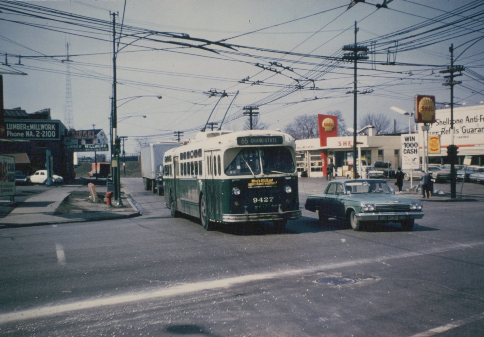 A green and cream trolleybus with the number 9427 on the front is driving on a city street, with a Shell gas station and other vehicles in the background.