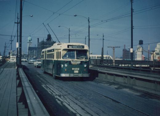 A green and cream Chicago Transit Authority trolleybus, number 9432, is shown on an elevated wooden bridge, with a line of 1950s cars behind and industrial buildings in the background under an overcast sky.