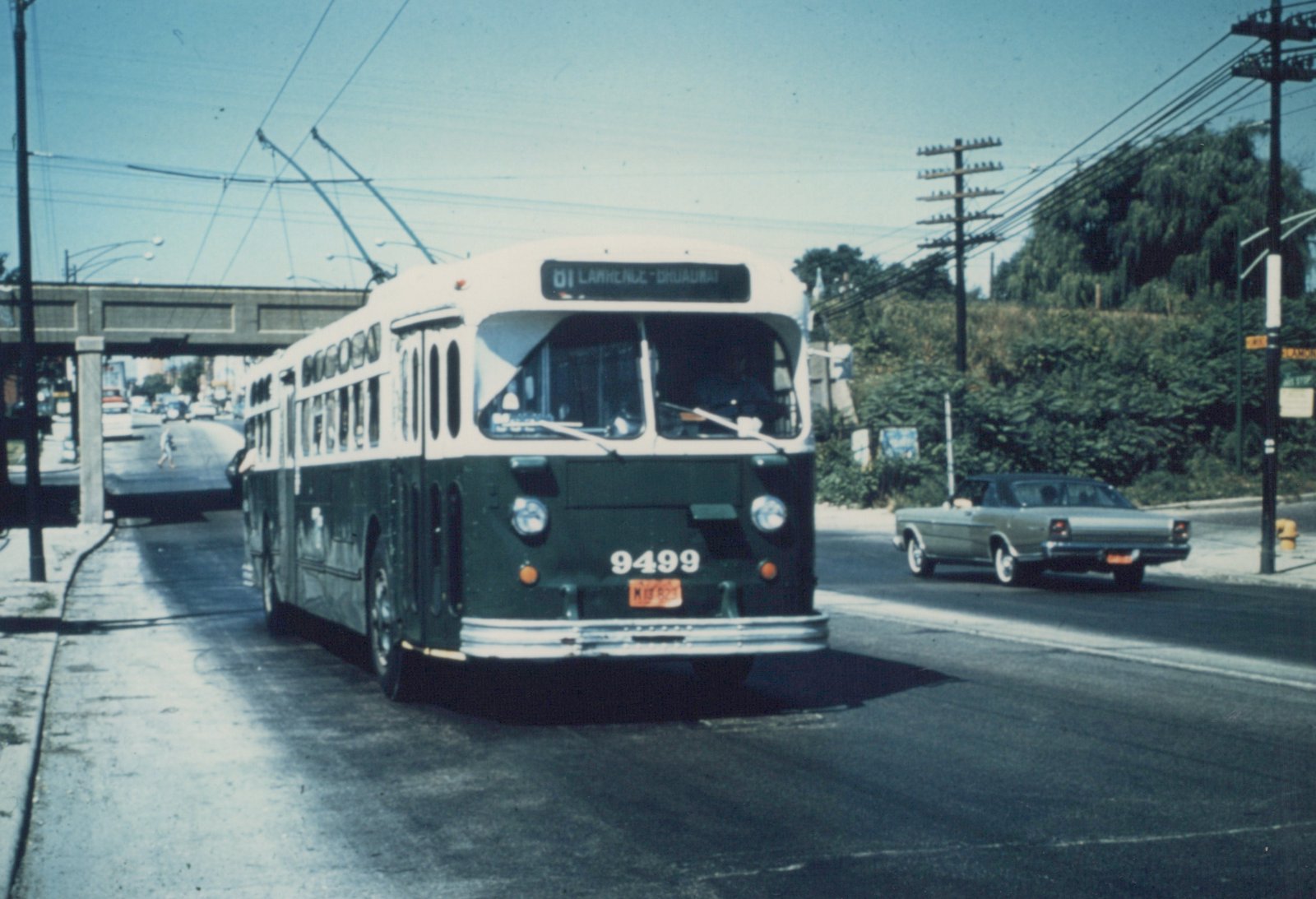 A green and white Chicago Transit Authority trolleybus on the 01 Lawrence route, with overhead wires and a 1960s sedan in the background, on a sunny street scene.