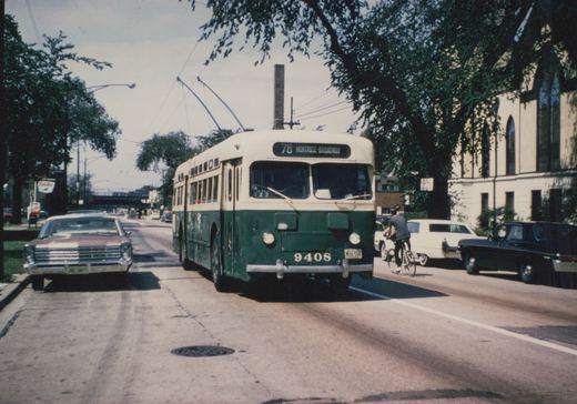 A green and cream trolleybus with the number 9408 and destination sign '78 MONTROSE-BROADWAY' is driving down a tree-lined street, sharing the road with cars and a cyclist. Overhead wires power the bus.