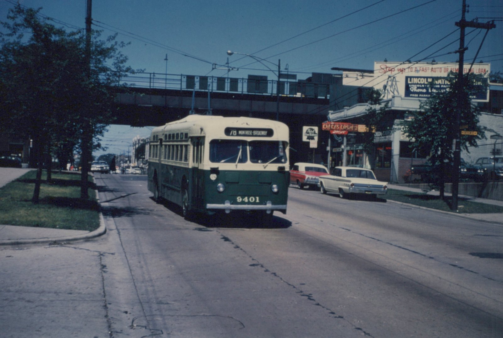 A green and cream Chicago Transit Authority trolleybus numbered 9401 is driving down a street under an elevated train track, with 1960s cars parked nearby and businesses in the background.
