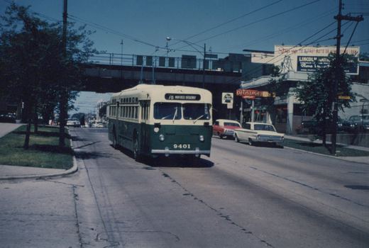 A green and cream Chicago Transit Authority trolleybus numbered 9401 is driving down a street under an elevated train track, with 1960s cars parked nearby and businesses in the background.