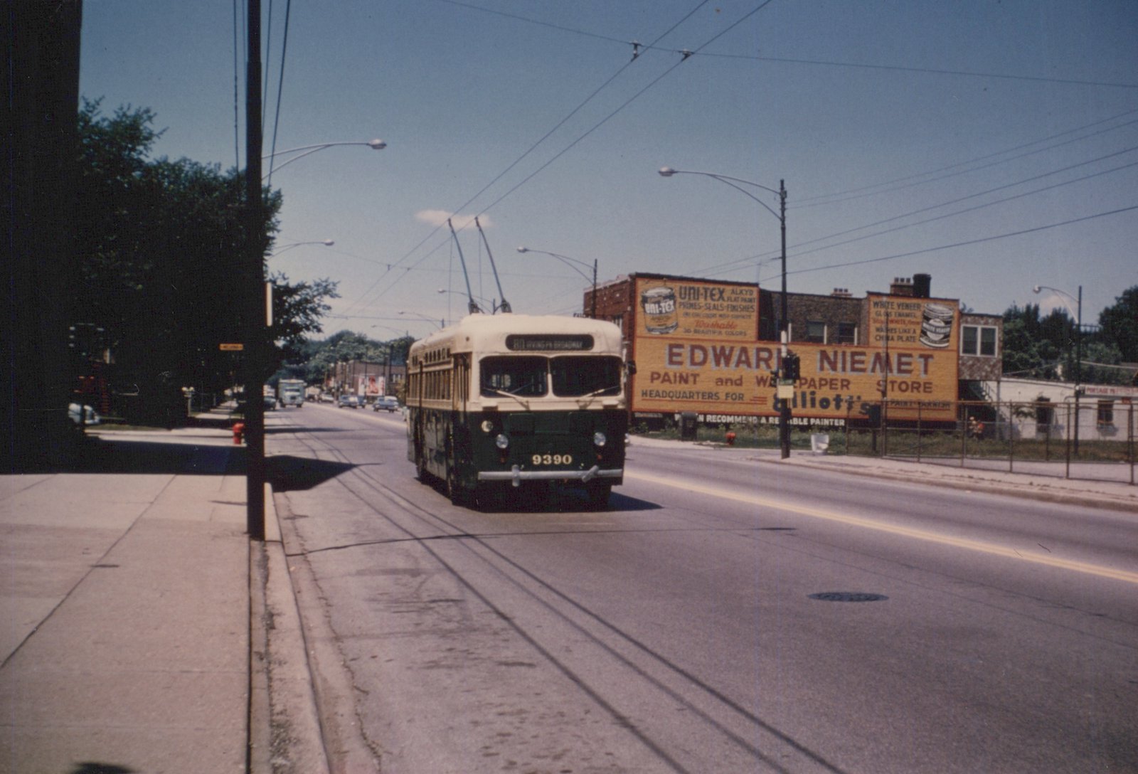 A green and white trolleybus with the number 9390 and route sign '80 IRVING PK BROADWAY' is driving down a street with visible tracks and overhead wires, passing by a large 'Edward Niemet Paint and Wallpaper Store' billboard. The scene is set on a clear, sunny day.