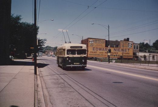 A green and white trolleybus with the number 9390 and route sign '80 IRVING PK BROADWAY' is driving down a street with visible tracks and overhead wires, passing by a large 'Edward Niemet Paint and Wallpaper Store' billboard. The scene is set on a clear, sunny day.