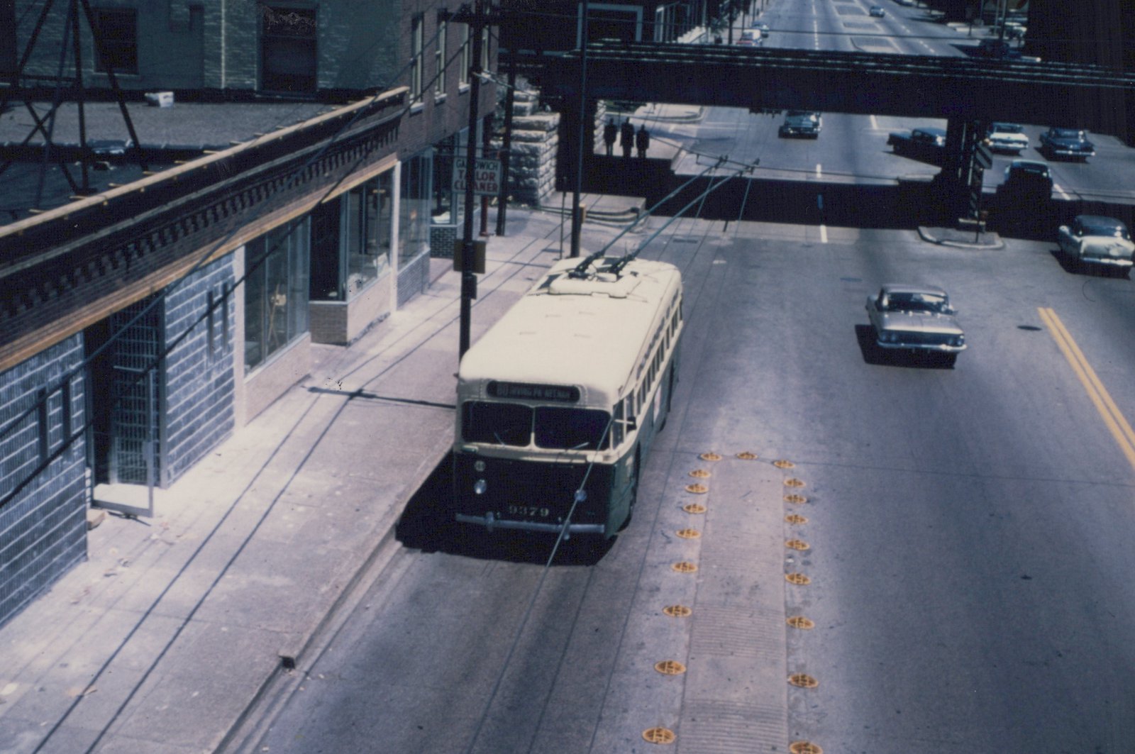 High-angle view of a green and white Chicago Transit Authority trolleybus, number 9379, on a city street with overhead wires, an elevated structure, and vintage 1960s cars.