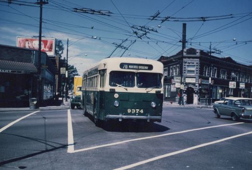 A green and cream CTA trolleybus, number 9374, is turning at an intersection under overhead wires, with buildings and a vintage car in the background.