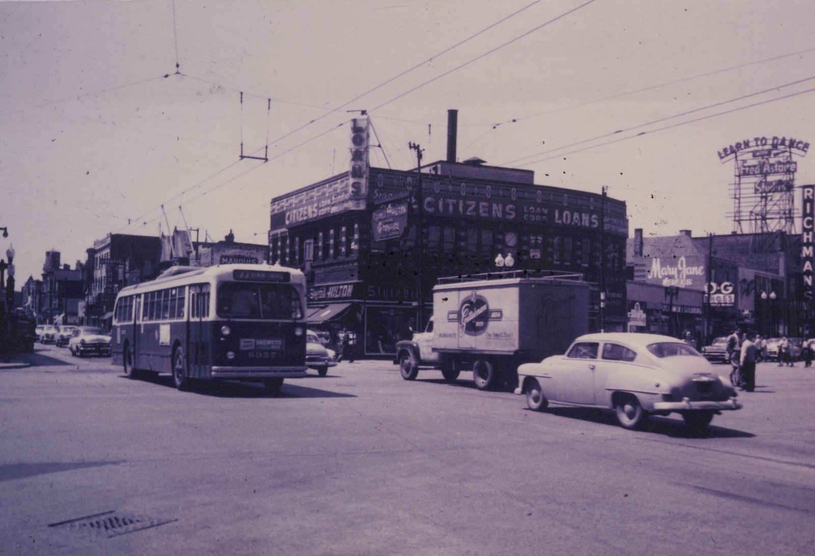 A vintage trolleybus travels down a busy street in Chicago, with storefronts like Citizens Loan Corp and a Fred Astaire dance studio sign in the background.