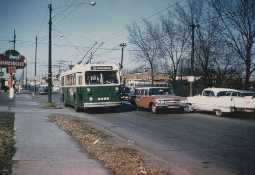 A green and cream trolleybus with the number 9355 and destination sign reading '85 CENTRAL-HARRISON' is on a city street lined with vintage cars and bare trees, with a 'Henry's Hamburgers' sign visible on the left.
