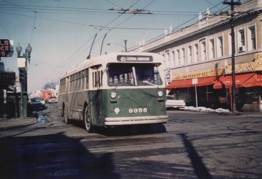 A green and cream CTA trolleybus numbered 9355 on route 85 Central-Harrison travels down a sunny, snow-dusted street lined with storefronts and power lines.