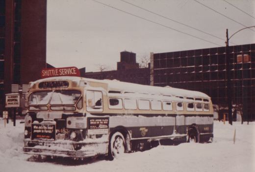 A snow-covered bus with 'Shuttle Service' on its roof sign and the number 5482 on its front and rear, parked on a snowy city street.