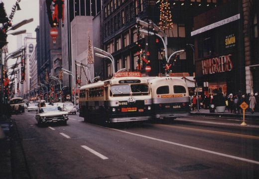Two cream and green Chicago Transit Authority trolleybuses with overhead wires drive down a busy city street decorated for Christmas, with Walgreens and other storefronts visible in the background.
