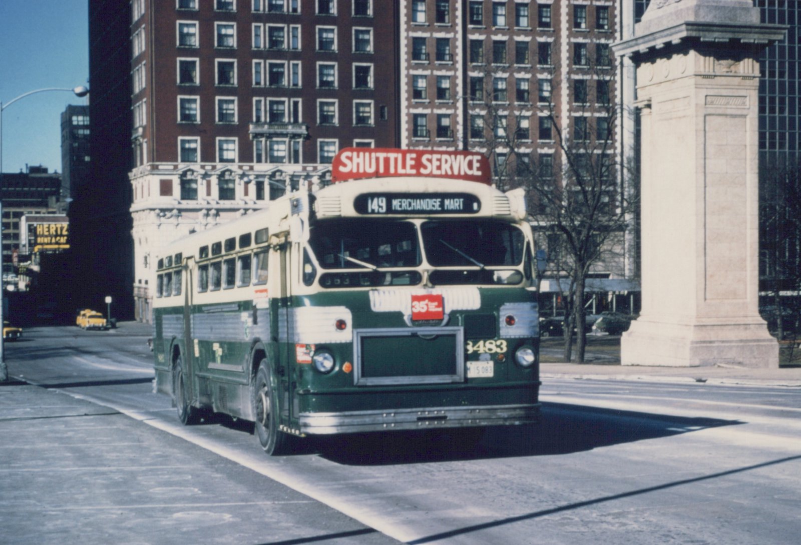 A green and cream CTA bus with the number 3483 on its front, displaying route 149 to Merchandise Mart, parked on a city street near a large building with a Hertz sign.
