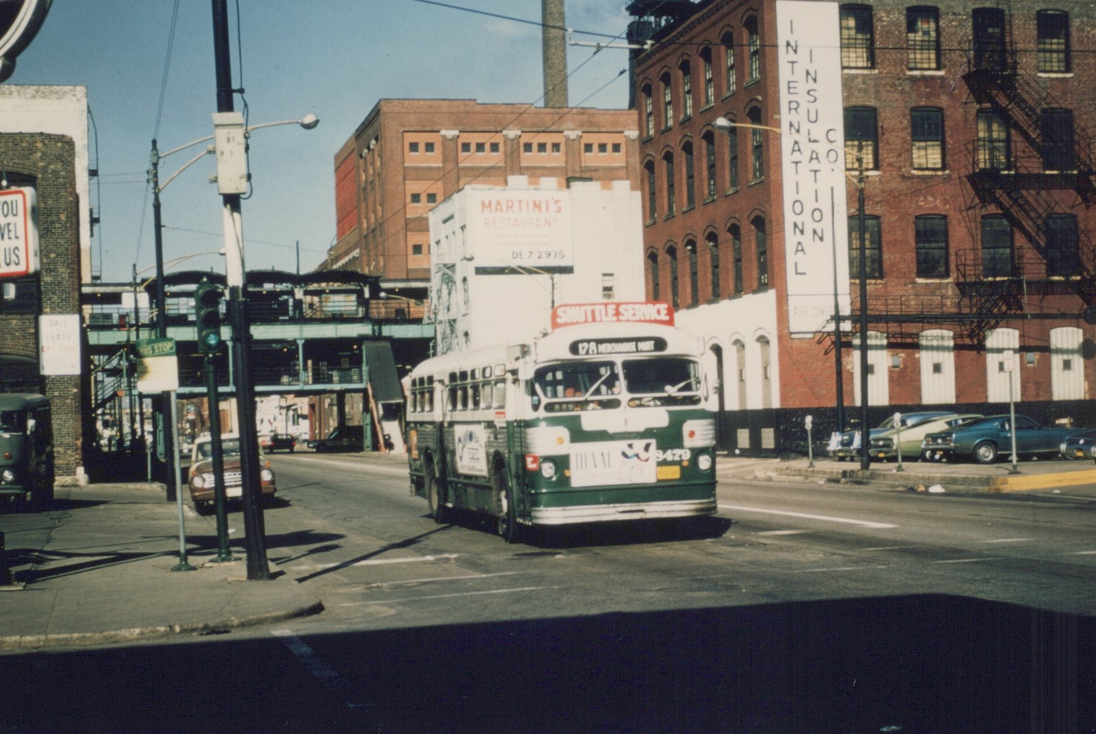 A green and cream CTA bus is stopped at an intersection with a 'Shuttle Service' sign, surrounded by brick buildings and parked cars under a clear sky.