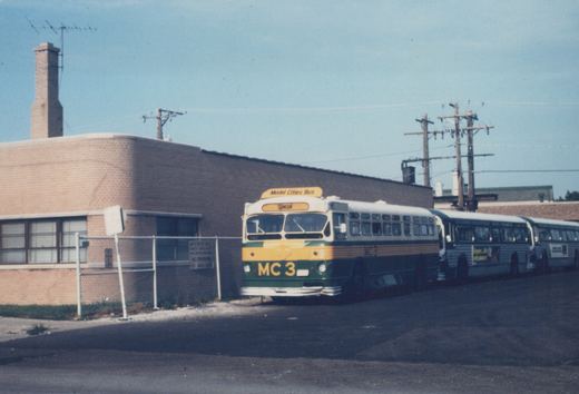 A vintage green, yellow, and cream bus with 'MC 3' on the front is parked on an asphalt lot beside a brick building, with two other buses lined up behind it.