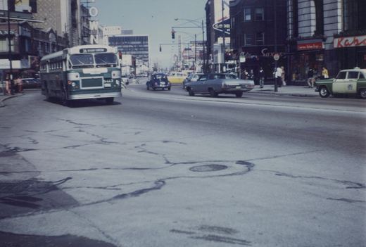 A vintage Chicago Transit Authority bus, number 8413, is driving down a busy city street lined with storefronts and cars from the 1960s or 1970s.