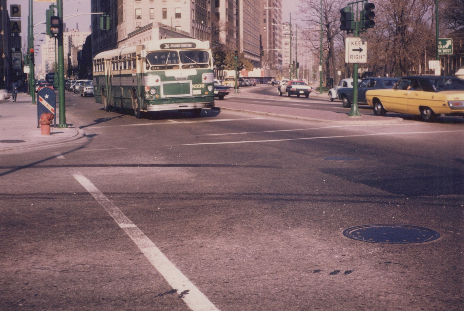 A green and cream CTA bus, number 8336 on route 36 Broadway-Downtown, turns at a city street intersection lined with 1970s cars and tall buildings.