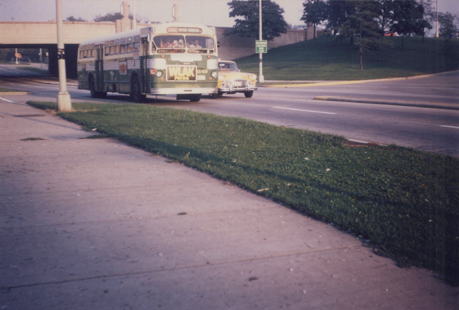 A green and cream Chicago Transit Authority bus numbered 6149 is driving on a road near a grassy median and a concrete underpass; a yellow taxi is behind it, and a street sign reads 'Lake Shore Drive'.