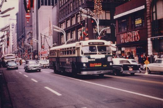 A green and cream trolleybus with the number 8073 on its front is driving down a decorated downtown street lined with tall buildings and vintage cars, with pedestrians walking on the sidewalk.