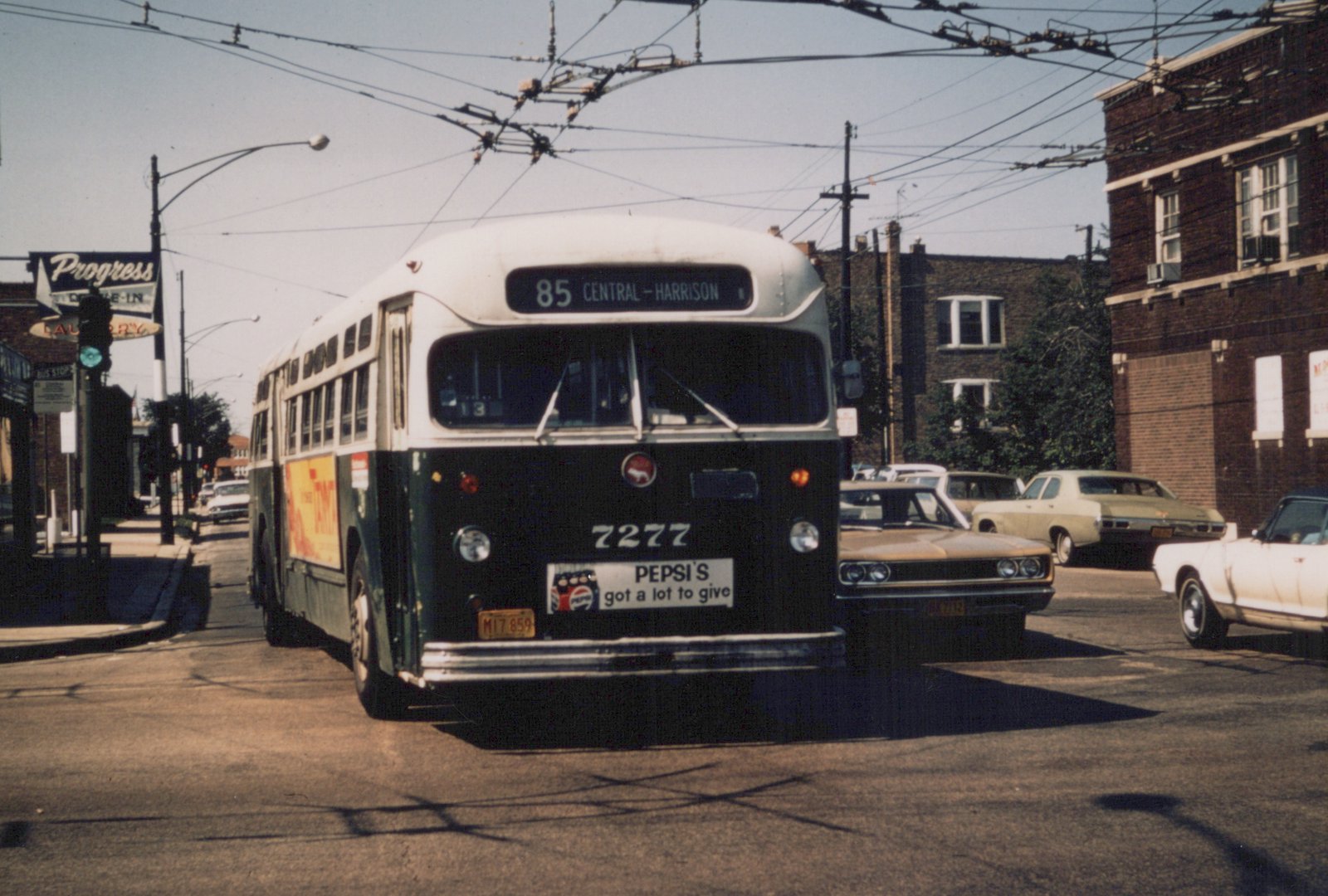 A front three-quarter view of a dark green and white CTA bus, number 7277, on a sunny street with cars and brick buildings in the background.