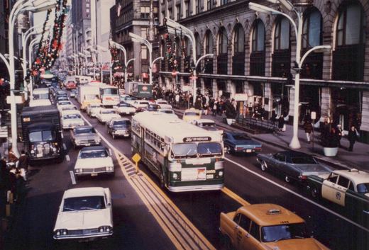 A green and white CTA bus driving down a crowded city street lined with decorated lampposts and vintage cars, with pedestrians on the sidewalk.