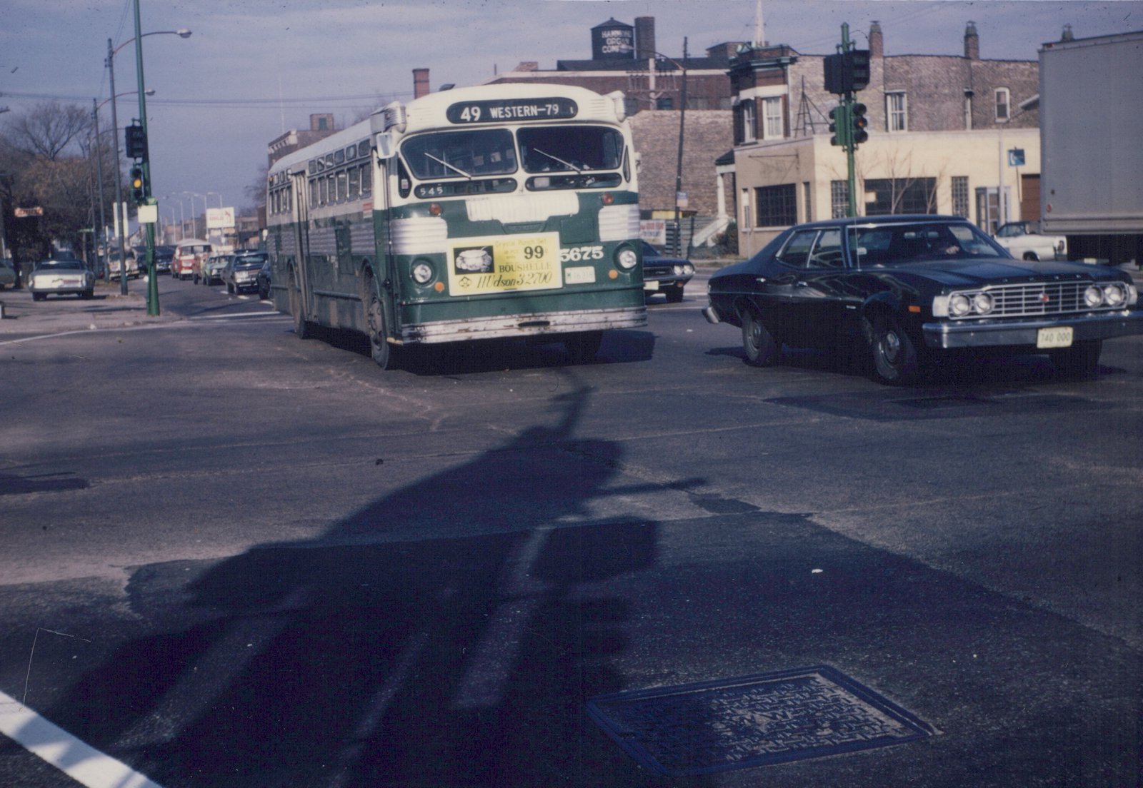 A green and cream Chicago Transit Authority motor bus is turning at a street intersection with other cars and buildings in the background on a clear day.