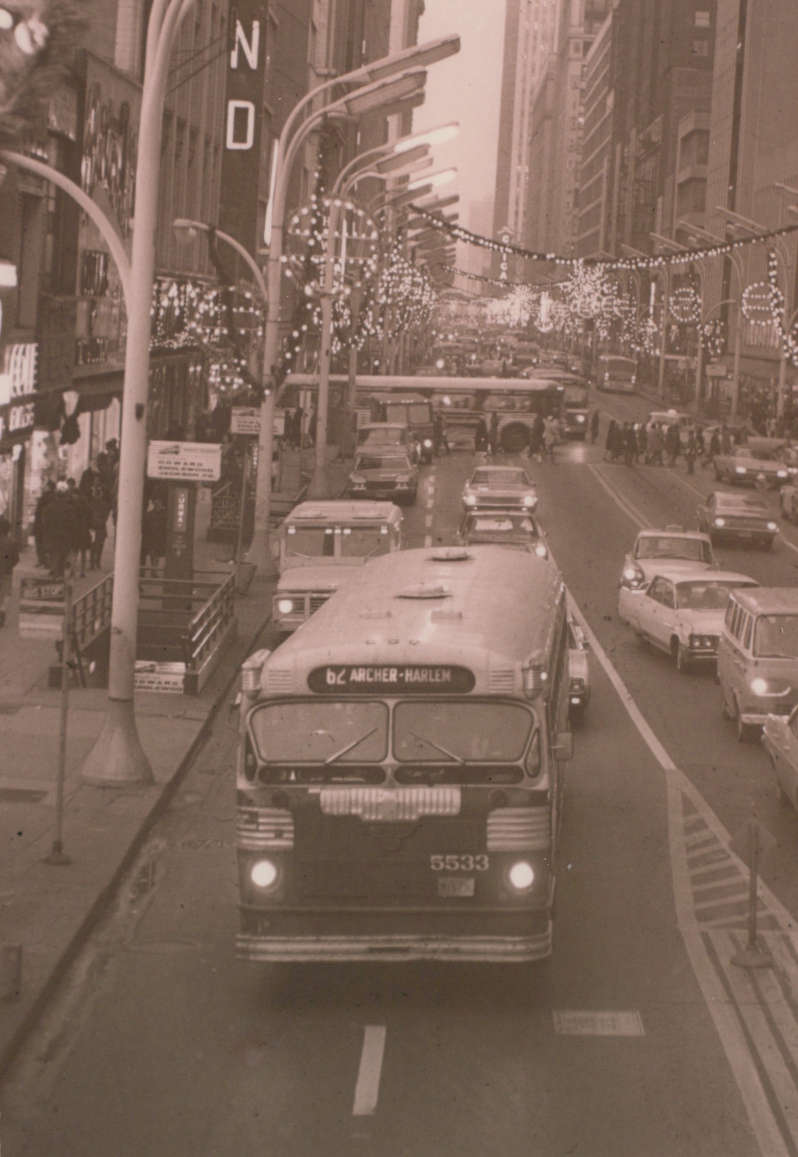 A black and white photo of a Chicago Transit Authority bus, number 5533, on the 62 Archer-Harlem route, driving through a crowded city street lined with decorated trees and tall buildings.