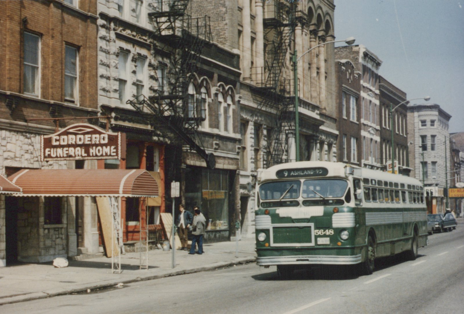 A green and white Chicago Transit Authority bus numbered 5648 is driving down a city street with a '9 Ashland-95' destination sign, passing a funeral home and two people on the sidewalk.