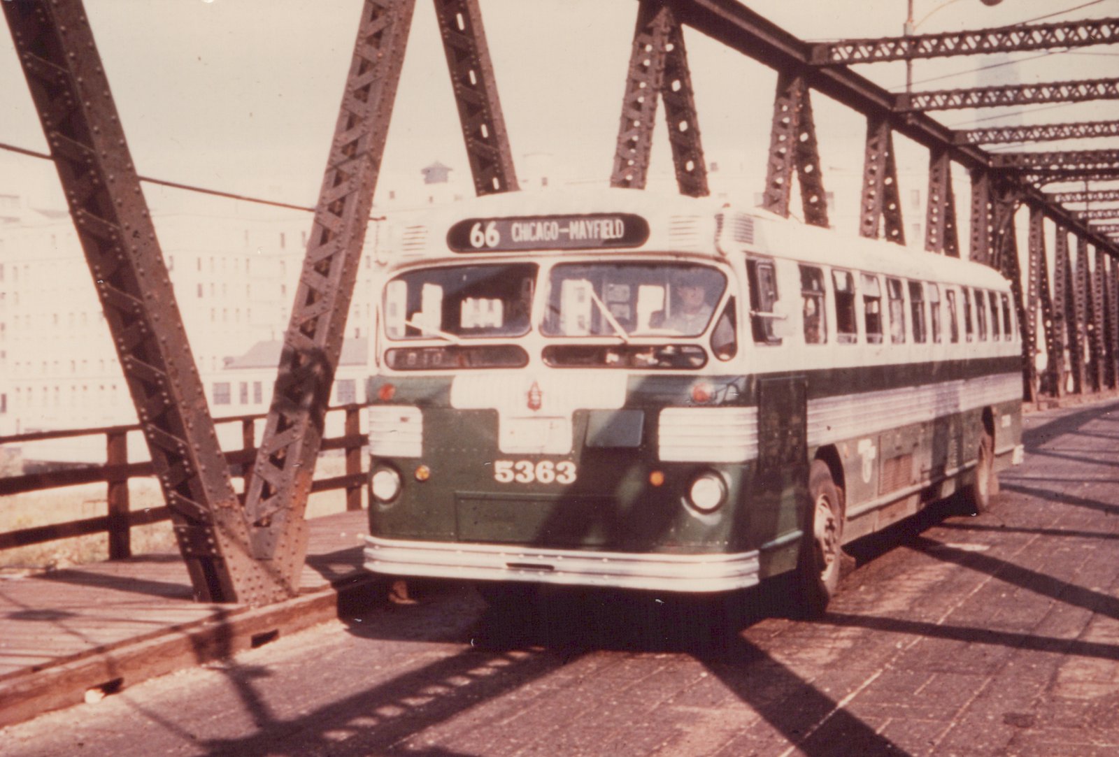 A green and white CTA motor bus, number 5363, on route 66 to Chicago-Mayfield, is seen from a three-quarter front view as it crosses a steel truss bridge on a sunny day, with its shadow cast on the pavement.