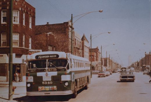 A green and white CTA bus with the number 5340 and a sign for route 70 Division-Austin is stopped on a city street lined with brick buildings and vintage cars.