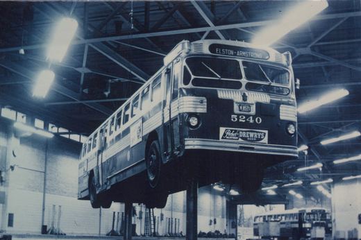 A black and white photo of a Chicago Transit Authority bus, number 5240, elevated on a lift inside a large garage, with its destination sign reading 'ELSTON-ARMSTRONG' and an ad for Drewry's beer on the front.