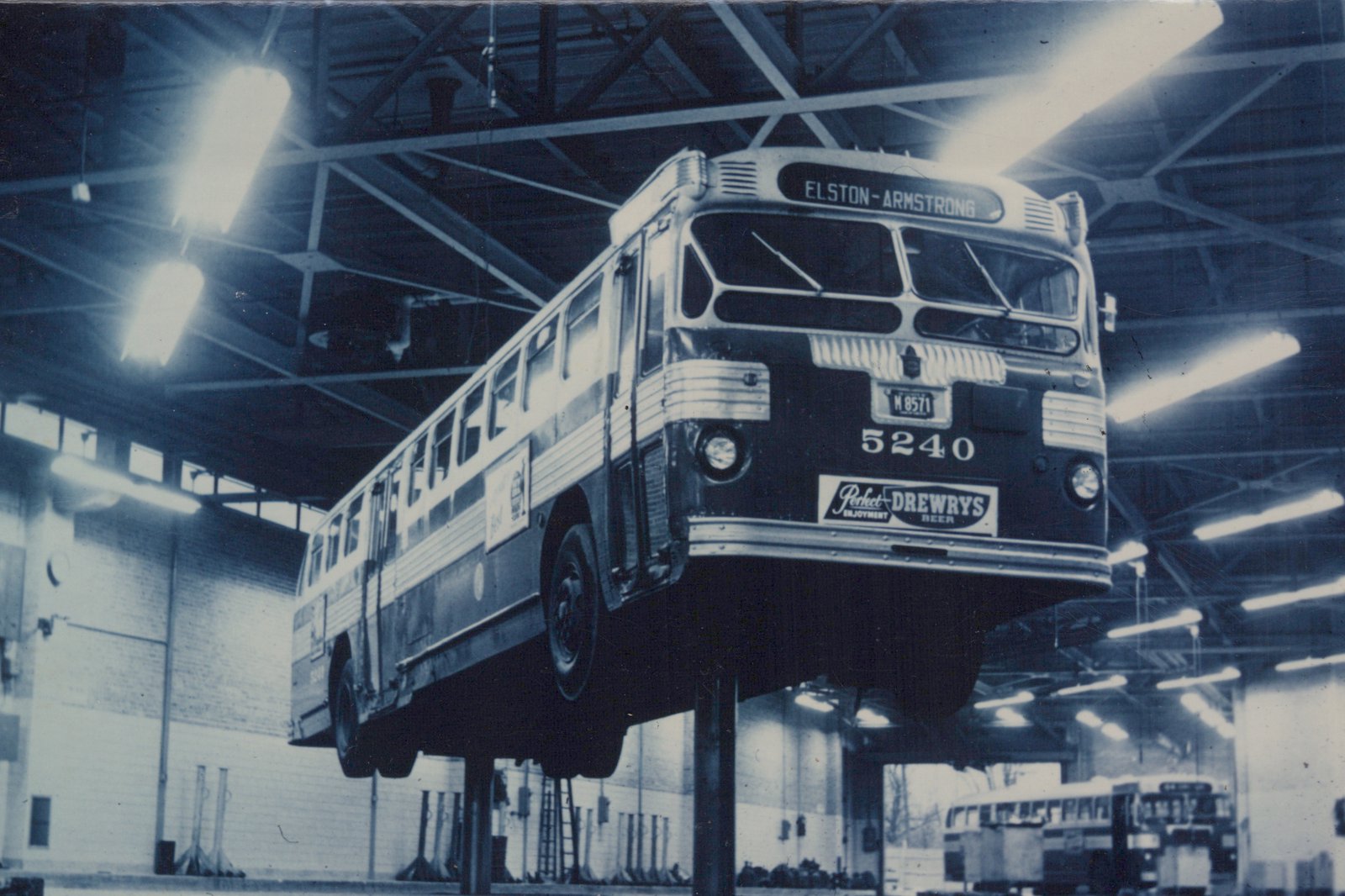 A black and white photo of a Chicago Transit Authority bus, number 5240, elevated on a lift inside a large garage, with its destination sign reading 'ELSTON-ARMSTRONG' and an ad for Drewry's beer on the front.