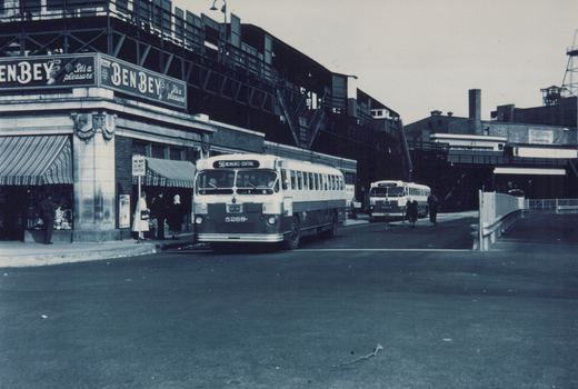 Black and white photo of two white Chicago Transit Authority buses on a city street under an elevated train track, with pedestrians and storefronts nearby.