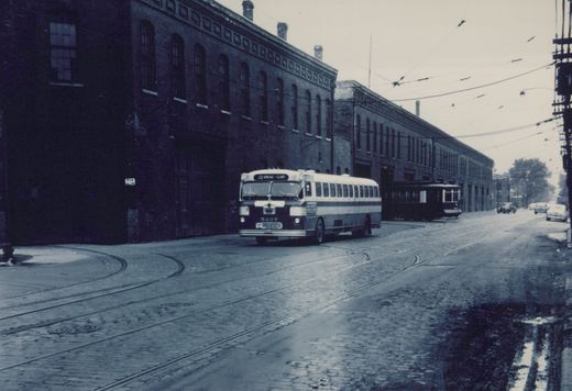 A black-and-white photo shows a 1950s CTA bus numbered 5207 on a cobblestone street with trolley tracks, a dark streetcar behind it, and brick buildings lining the road.
