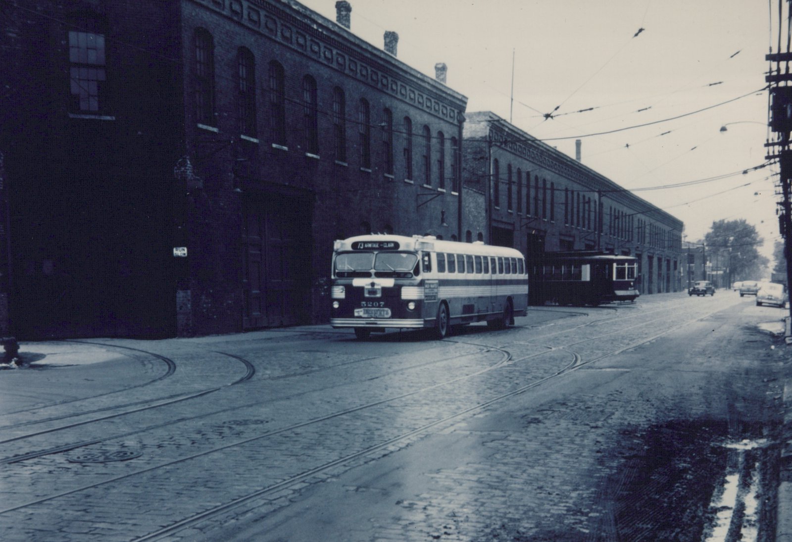 A black-and-white photo shows a 1950s CTA bus numbered 5207 on a cobblestone street with trolley tracks, a dark streetcar behind it, and brick buildings lining the road.
