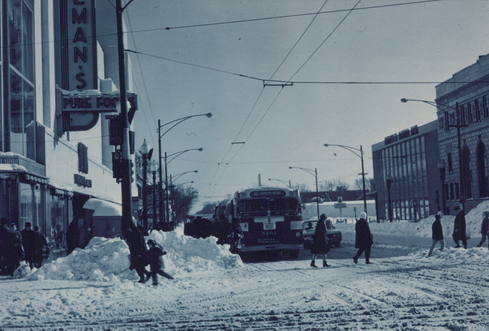 A black and white photo of a CTA trolleybus on a snowy street in Cicero, Illinois, with people walking nearby and storefronts lining the street.
