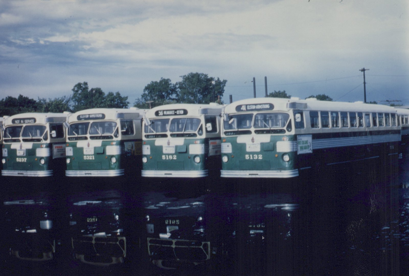 Four green and cream CTA buses, numbered 5132, 5182, 5321, and 5237, are parked side by side in a yard with their reflections visible in a puddle on the ground.
