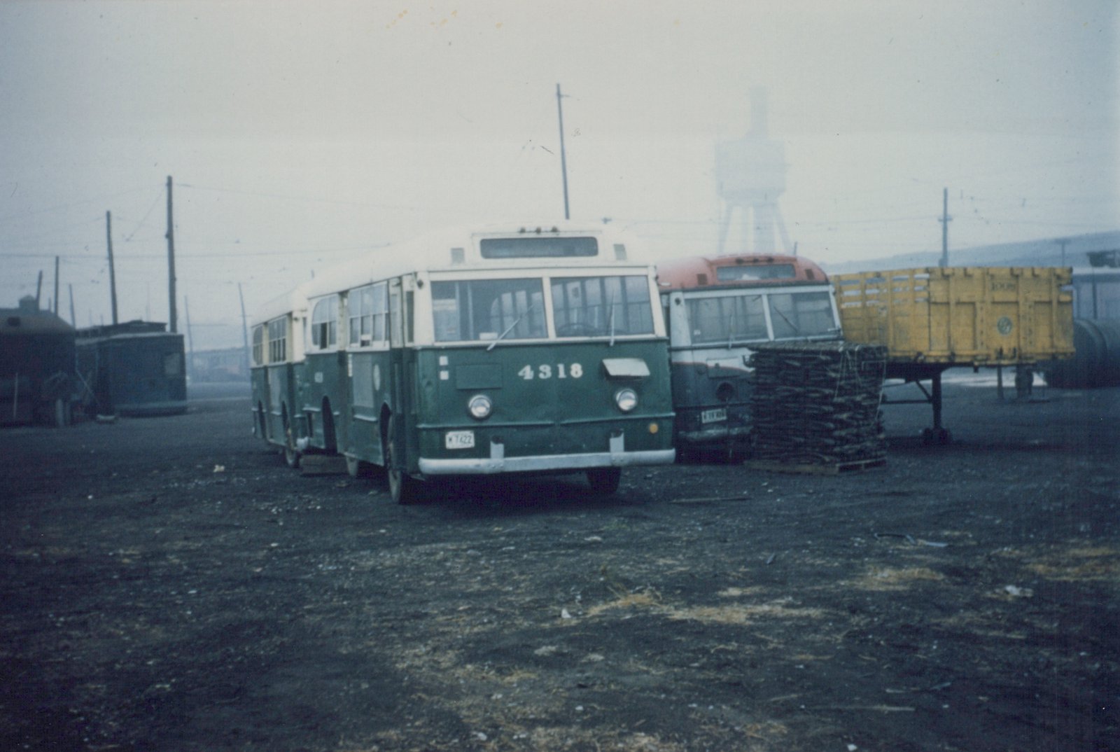 Three-quarter front view of a green and cream CTA bus numbered 4318 parked in a gravel yard under an overcast sky, with another bus and a yellow freight car visible behind it.