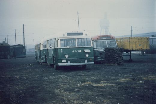 Three-quarter front view of a green and cream CTA bus numbered 4318 parked in a gravel yard under an overcast sky, with another bus and a yellow freight car visible behind it.